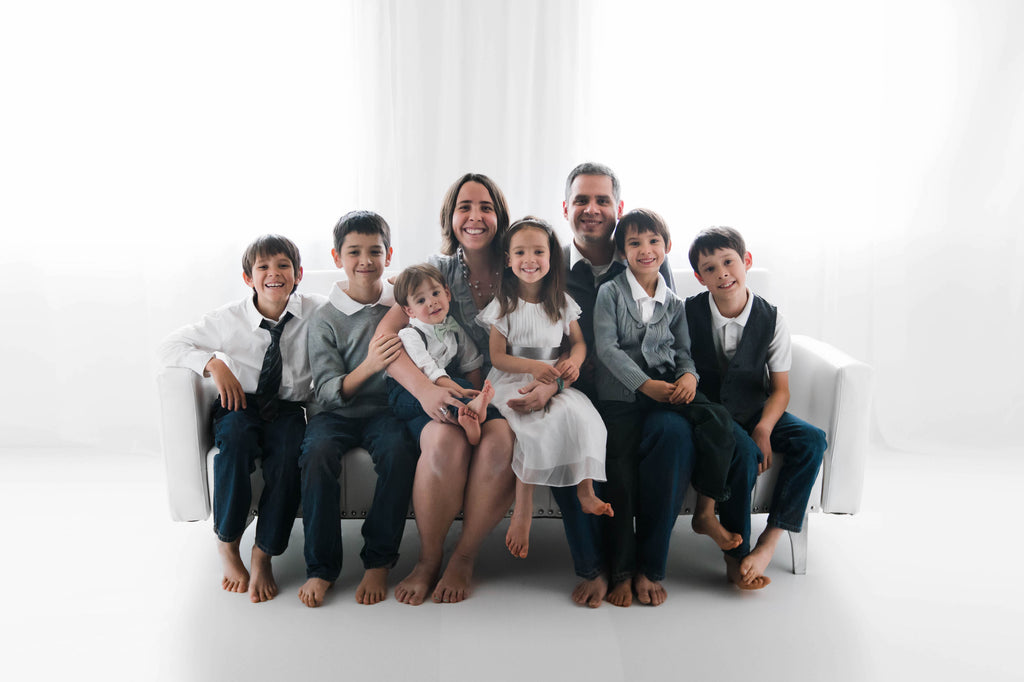 this is a family photo of the family that owns colorado baby. the family is sitting on a couch in an all white room in a photography studio. the mom is white with brown hair, the father is latino with black hair. there are six kids on the couch with them