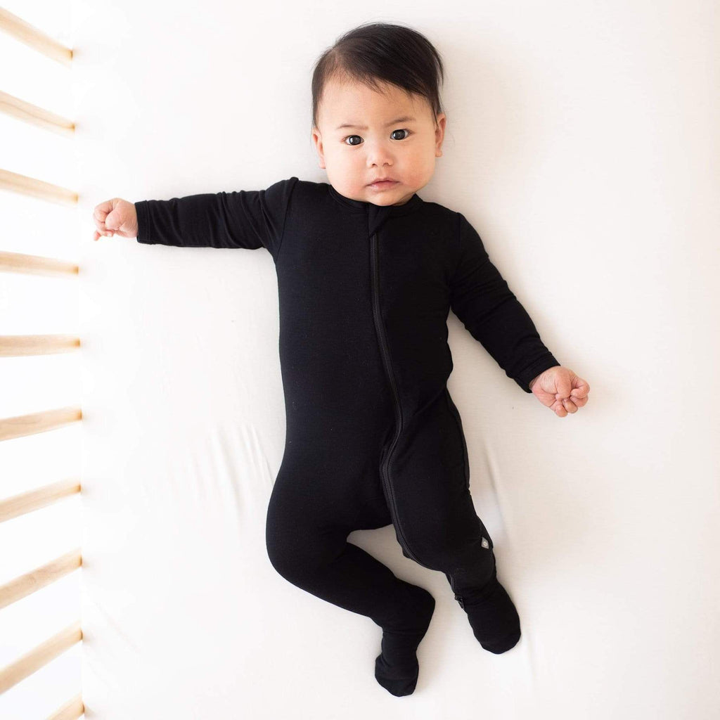 a six month old baby laying in a crib looking up at the camera while wearing a black kyte baby footie